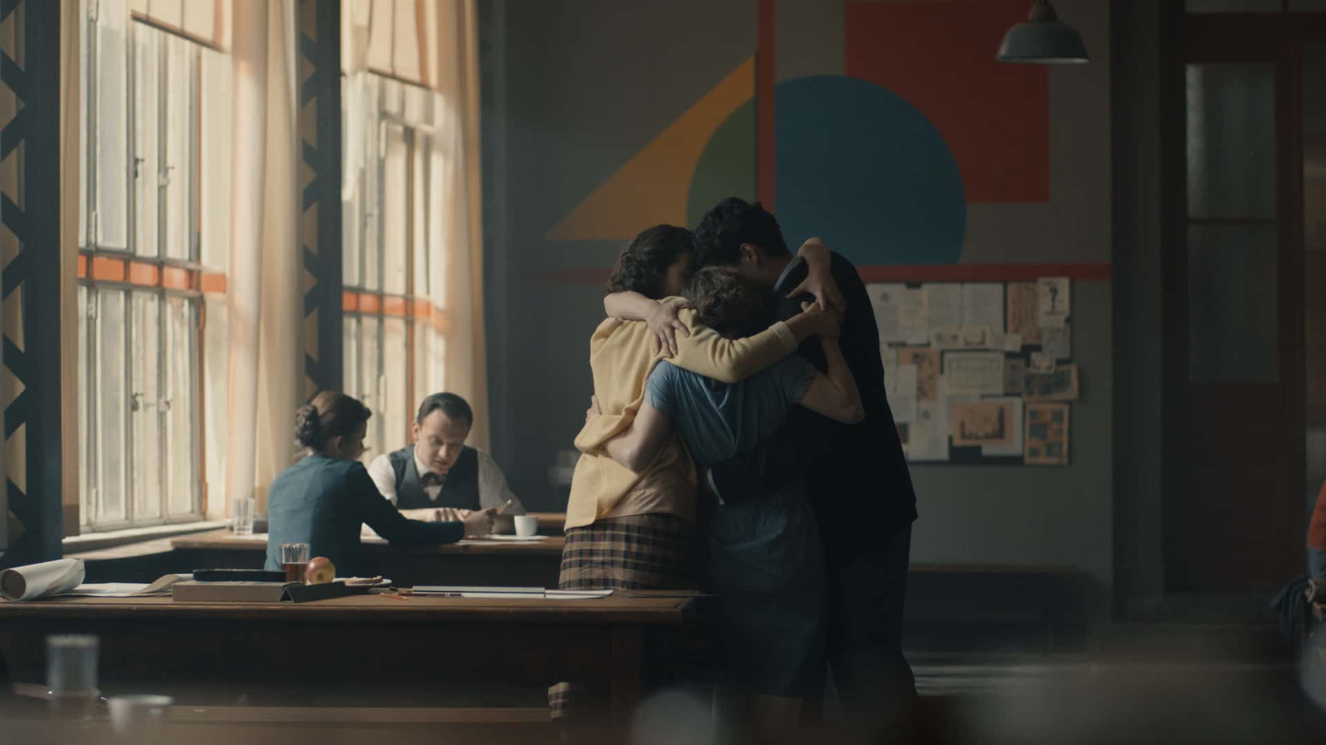 Filmszene. Im Vordergrund umarmen sich Lotte, Paul, Friedl und Anni in der Bauhaus Kantine. Im Hintergrund ist an der Wand ein gelbes Dreieck, ein rotes Quadrat und ein blauer Kreis.